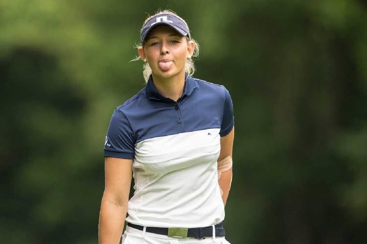 Kristine Pedersen with a tongue-out pose while wearing a white blue polo shirt, blue cap, black belt, and pants