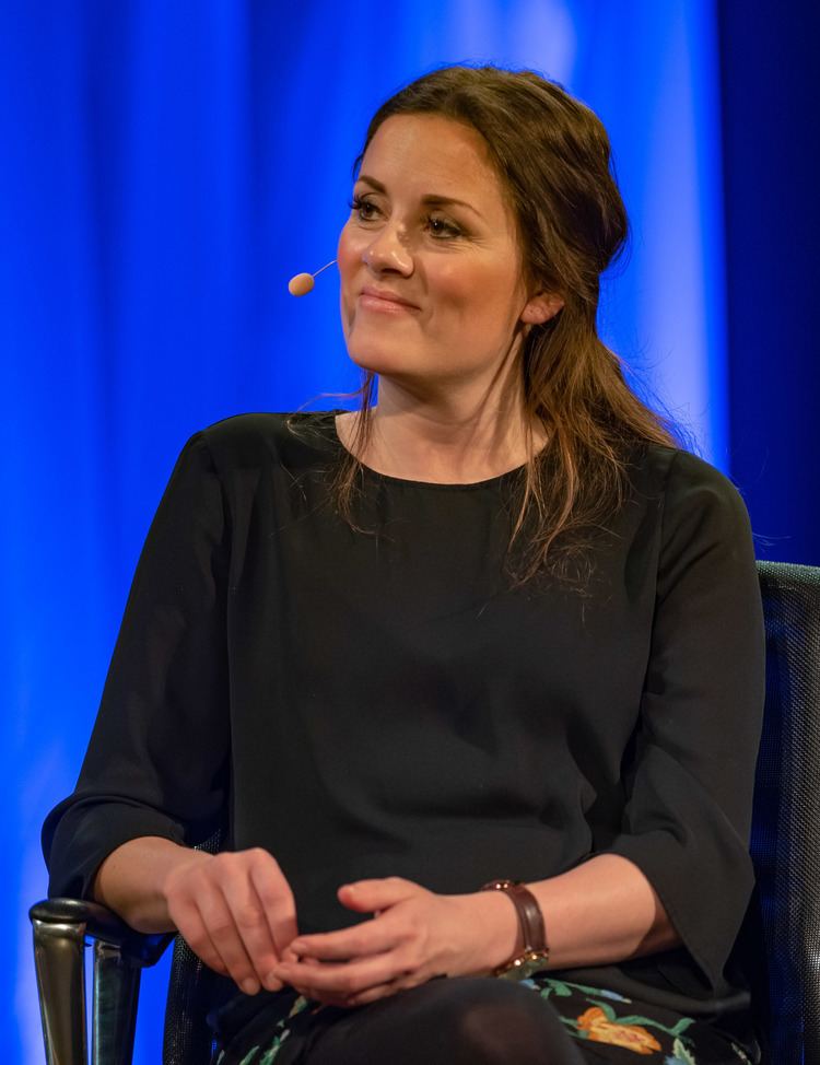 Kristine Riis smiling, with a wired microphone while sitting on a chair, wearing a brown watch, and a black dress.