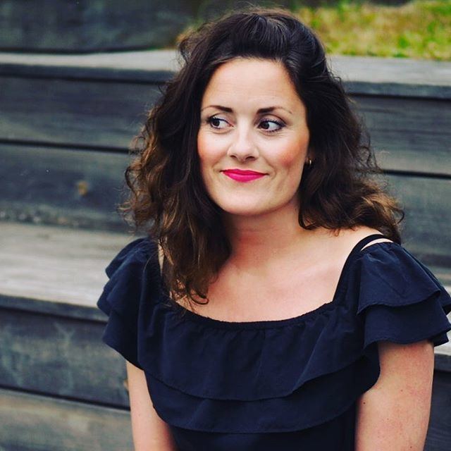 Kristine Riis smiling while looking on her right side, with curly hair, and wearing a dark blue blouse.