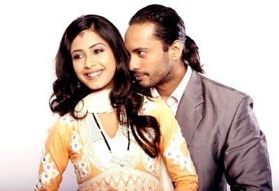 Dimple Jhangiani as Kanya/ Natasha smiling with long hair and wearing a yellow blouse and white scarf and necklace while being held from behind by Akashdeep Saigal as Ranbir Nanda having a ponytail hairstyle, a beard and mustache, and wearing a white shirt under a gray coat