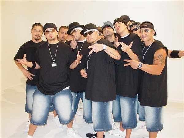 All the current members of Kumbia Kings posing with hand gestures and wearing black clothing and jean shorts.