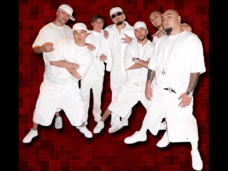 Seven members of Kumbia Kings posing with hand gestures and wearing white clothing and necklaces.