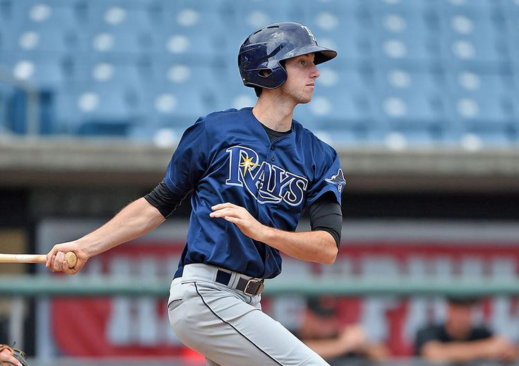 In a baseball field with blue chairs and banner at the back, Kyle Tucker is serious, hitting with his bat, right hand holding it from the back and left hand let go of the bat, has black hair, wearing a black helmet, a blue Rays Jersey over black dri-fit half sleeve, and a gray pants.