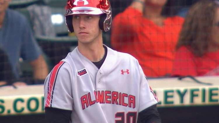In a baseball field Kyle Tucker is serious, playing, looking to his right, standing, wearing a black dri-fit long sleeve, under a gray All America Jersey and a red Under Armour helmet.