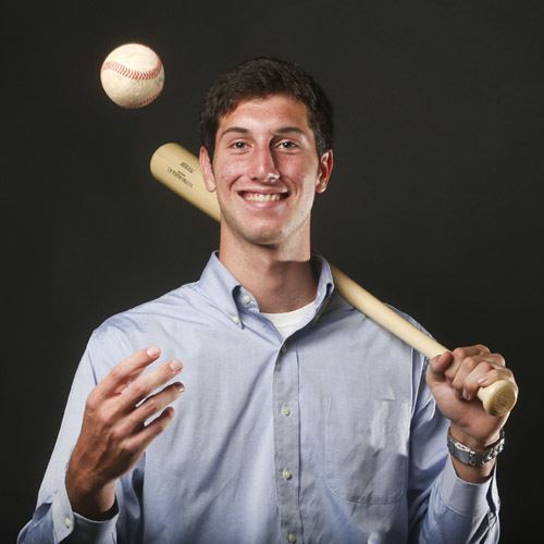 In a dark gray background Kyle Tucker is smiling, standing holding a bat with his left hand while playing with a ball with his right hand, has black hair wearing a white shirt under a sky blue long sleeve polo and a silver watch.