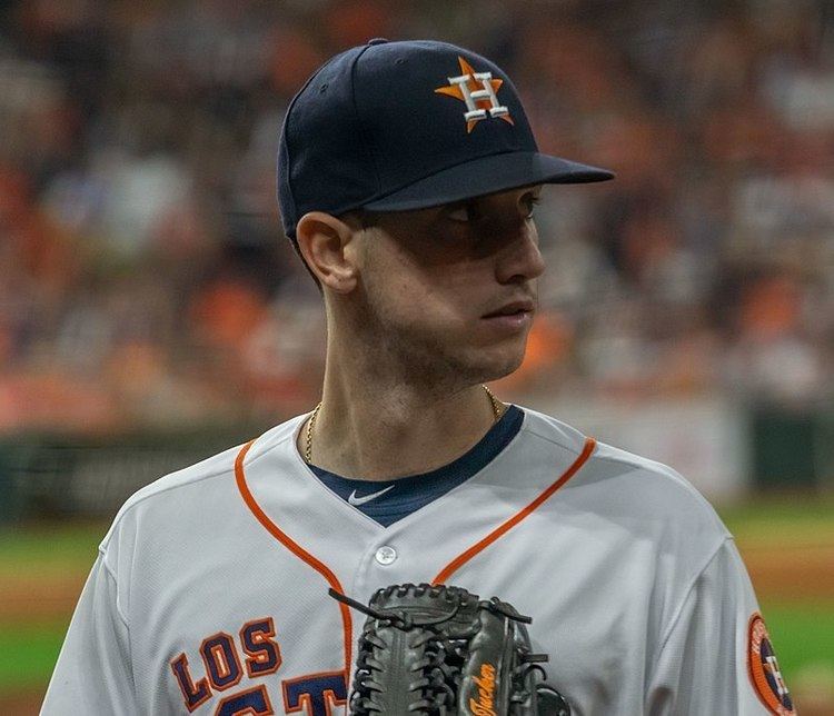 In a baseball field Kyle Tucker is serious, playing, looking to his left, wearing a black baseball cap with H in the middle of an orange star, a dark blue dri-fit Nike shirt under a gray Jersey and olive catcher's mitts.