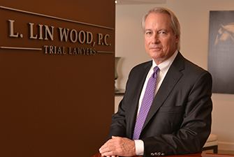 L. Lin Wood with a tight-lipped smile, with L. LIN WOOD, P.C. TRIAL LAWYERS in a brown wall, a white wall and a painting in the background. He is wearing a black coat over white long sleeves, and a purple necktie.