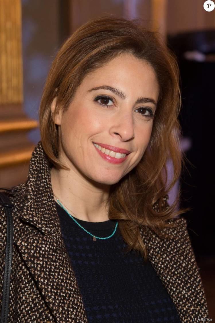 Léa Salamé smiling while wearing a brown coat, black blouse, and blue beaded necklace