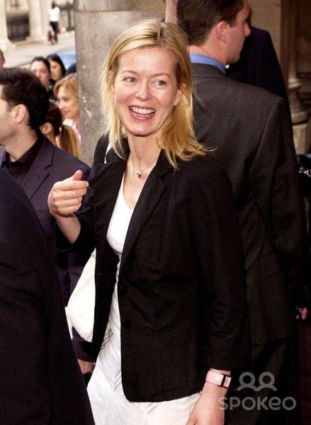 Lady Helen Taylor is smiling, has blonde hair, has a white bag on her right arm, is wearing a white wristwatch on her left hand, a black necklace, white dress under a black suit. She is surrounded by people.