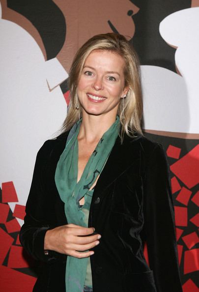 Lady Helen Taylor is smiling, has long blonde hair, her right hand in front of her stomach, wears silver earrings, a green cleavage showing top under a black suit.