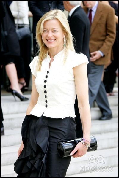Lady Helen Taylor is smiling, has blonde hair, her left hand holding a black purse, and wears silver earrings, a silver bracelet on her left hand, a white top with black buttons, and a black skirt. Behind her people are walking.