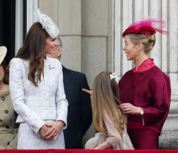 Catherine (left) Duchess of Cambridge is smiling and has brown way hair, both hands holding each other, wears a silver-black ring, a white hair clip, a white long-sleeve top, and a skirt. Estella Taylor (middle) has long blonde hair and wears a white clip and an off-white dress. Lady Helen Taylor (right) is smiling, mouth half opened, has blonde hair, left hand holding Estella’s hair, right hand poking Catherine, she is wearing silver earrings, a silver watch on her left hand, ring in left-hand finger a magenta hair clip, magenta long-sleeve top, and skirt. Behind Catherine on (left) is a woman, wearing an off-white hat and brown dress with black buttons, a man on the right has white hair and wears a black suit.