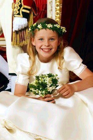 Margarita Armstrong-Jones is smiling, sitting, hands holding a small bouquet of flowers, has brown-blonde hair with flower crown on top, wearing a white dress, at the back is a royal guard wearing a red royal guard uniform a white gloves, white pants, white socks and black shoes,