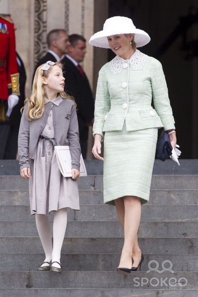 From left, is Lady Margarita Armstrong-Jones is serious, walking down the stairs, has blonde hair with gray ribbon on clip, wearing gray dress under a dark-gray coat, white socks and black shoes, at the right is Lady Serena Armstrong-Jones is smiling, walking down the stairs left hand holding a black small bag and white gloves, has blonde hair, wearing a white floppy hat, white earrings, cream green, long sleeve coat and skirt. A the back from left, a man, standing wearing a royal guard uniform, and white gloves, 2nd from left a man is serious standing, has black bald hair, wearing a white polo and black coat, 3rd from left a man is serious standing, has black hair, wearing a white polo and black coat,