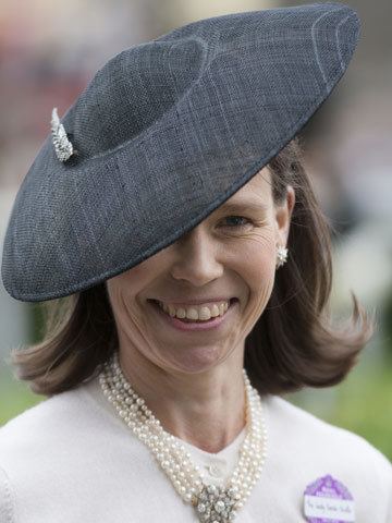 Lady Sarah Chatto is smiling, has black hair, wearing a black hat with a pin, silver earrings, and a white pearl necklace, and wearing a white top with a nametag.