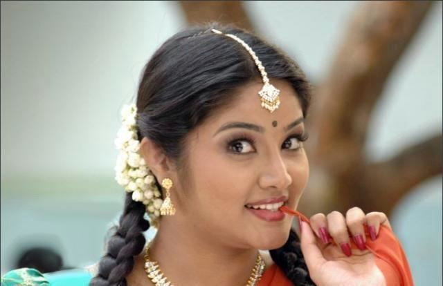 Lakshana smiling while biting her orange dupatta and wearing an aqua blue and gold dress, necklace, earrings, and hair accessory