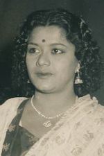 Lalitha smiling with curly hair and with a red mark on her forehead while wearing  a necklace and dangling earrings