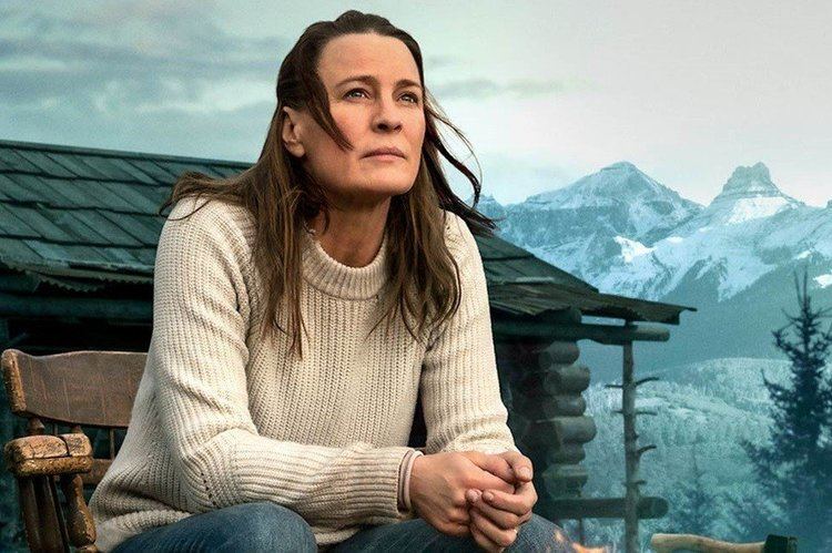 Robin Wright sitting on a chair, looking serious while wearing a white knitted jacket and jeans