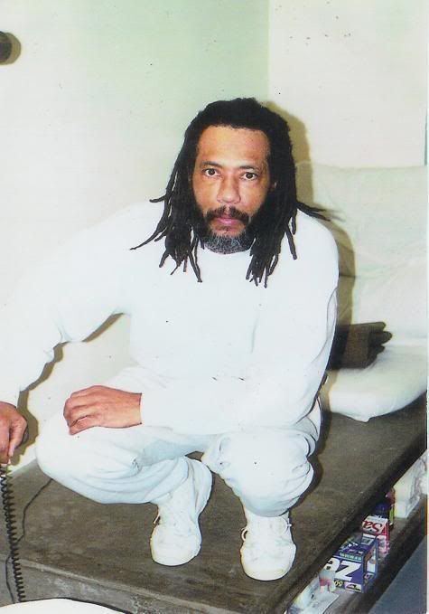 Larry Hoover sitting on a wooden bed while wearing a white sweatshirt, white pants, and white shoes