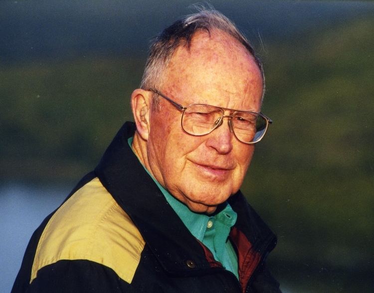 Larry Linville smiling while wearing a blue shirt under a colored a colored jacket and eyeglasses
