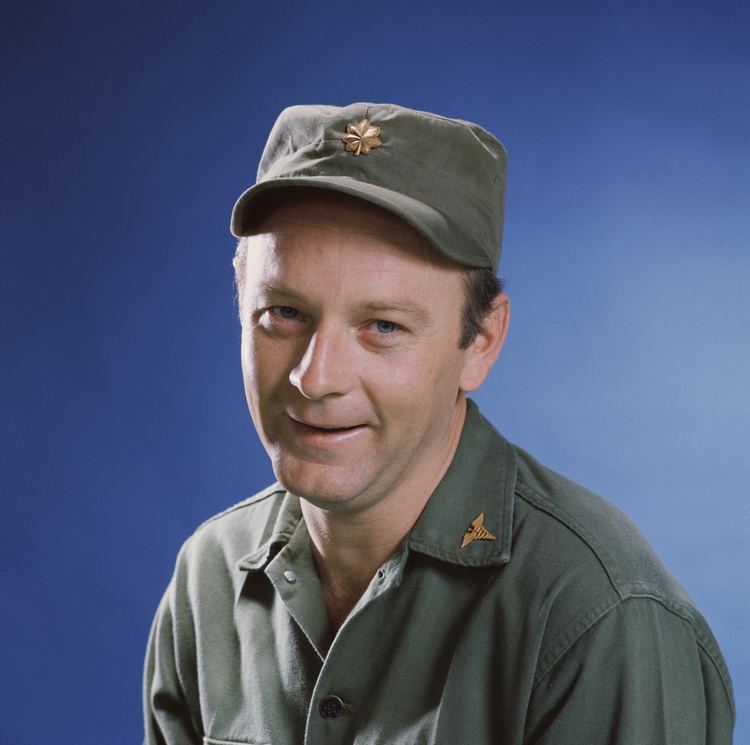 Larry Linville smiling in a green shirt and wearing a cap