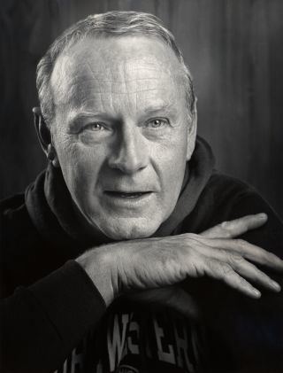 Larry Linville smiling with wrinkles on the forehead and hands below his chin while wearing a jacket