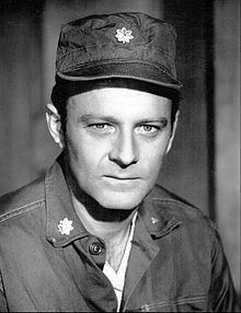 Larry Linville smiling and wearing a shirt and a cap