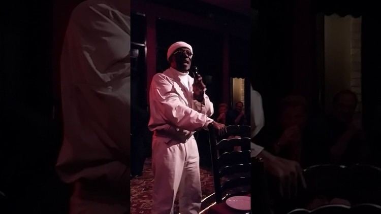 Larry McCoy is serious, standing while singing, right hand holding the brown chair, while left hand holding a mic, wearing a white bonnet black sunglasses white turtleneck under a white jacket and white pants.