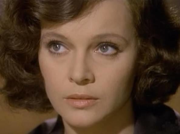 Laura Antonelli is looking serious while looking afar in Luigi Comencini's film, "My God, how did I fall so low?"(1974) screenshot. She has short curly hair, wearing a black collared blouse.