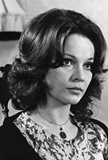 Laura Antonelli looking serious with short curly hair, wearing a pendant necklace, a scarf over her shoulders, and a printed blouse.