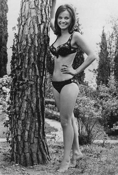 Laura Antonelli smiling while standing tiptoed, leaning her right elbow on a tree and her left hand on her waist has short hair with bushes and trees in the background. She is wearing a black printed two-piece bikini with visible cleavage.