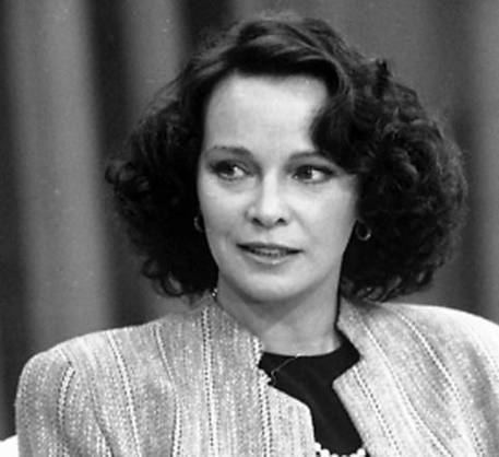 Laura Antonelli smiling while talking to someone having short perm-curl hair with a pair of hoop earrings and a curtain in the background. She is wearing earrings and a pearl-beaded necklace over a black inner blouse, and a striped coat.