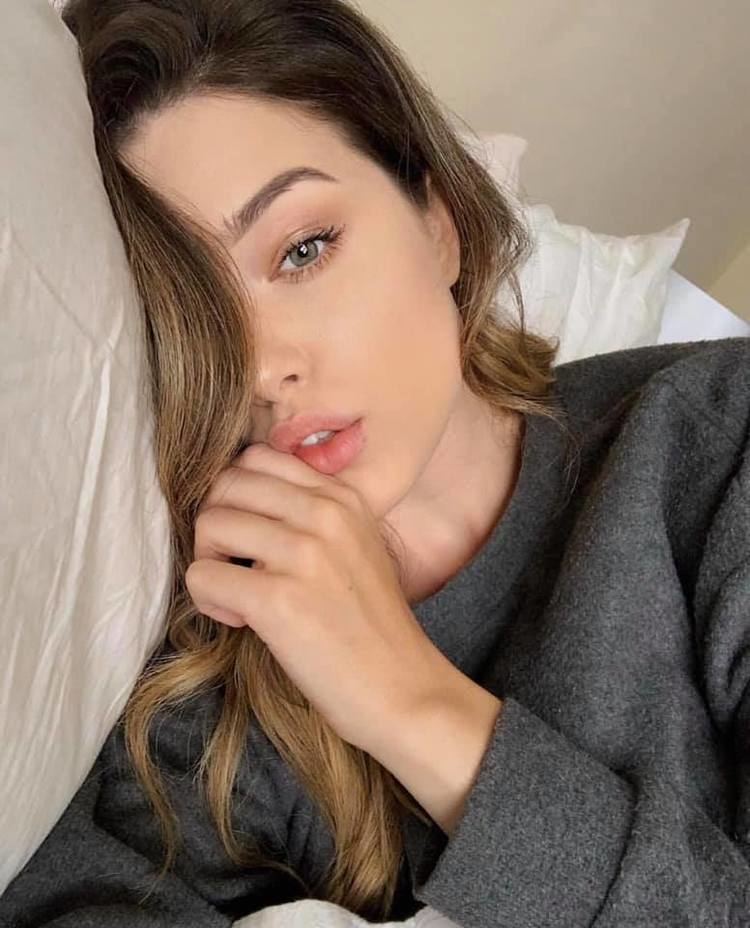 Lauren Summer with a serious face while lying on a bed, with wavy blonde hair, and wearing a gray sweatshirt.