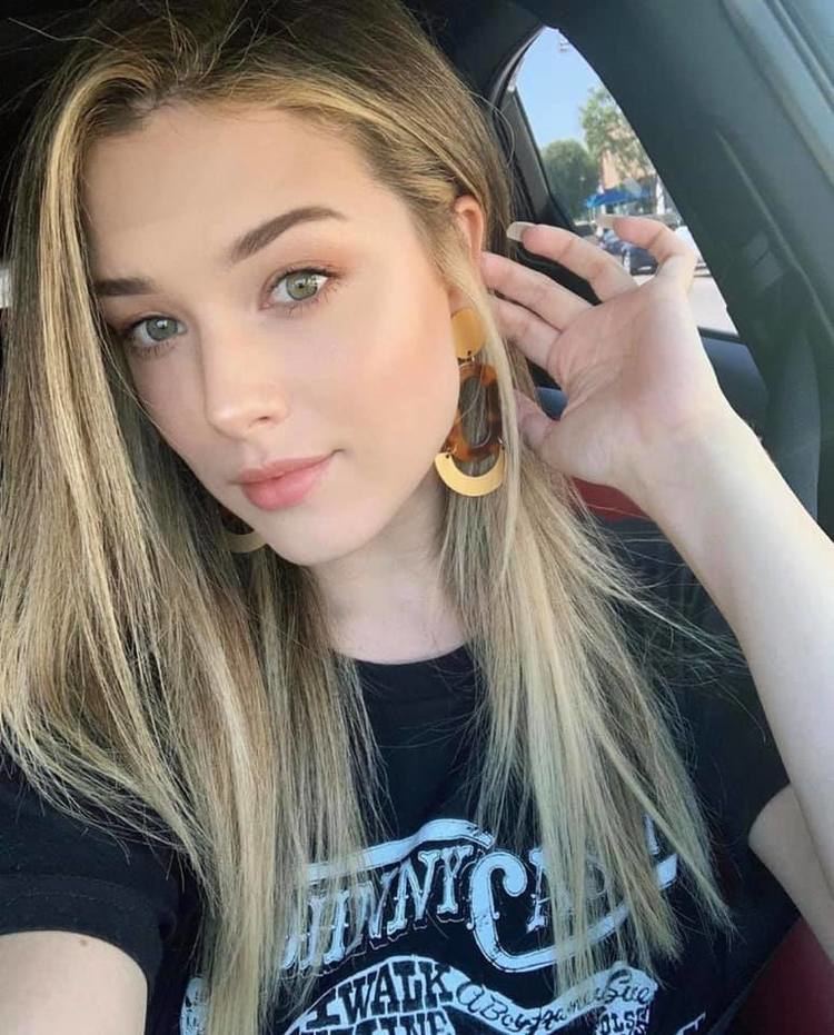 Lauren Summer with a serious face while holding her ear, with long blonde hair, wearing earrings and a black shirt with a white print.
