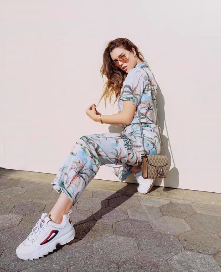 Lauren Summer with a fierce look while doing an extended leg pose, with wavy blonde hair, wearing sunglasses, a bracelet, white shoes, and a multi-colored floral top and pants.