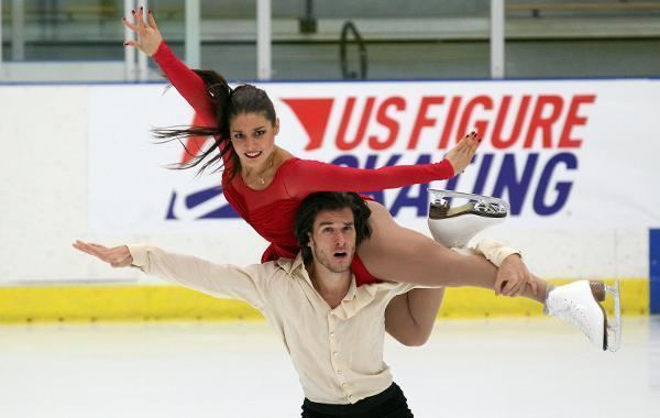Laurence Fournier Beaudry is on Nikolaj Sorensen shoulder, during a figure skate dance performance. Laurence wearing a long sleeve short red dress with her figure skate shoes while Nikolaj wearing a white long sleeve shirt and black pants