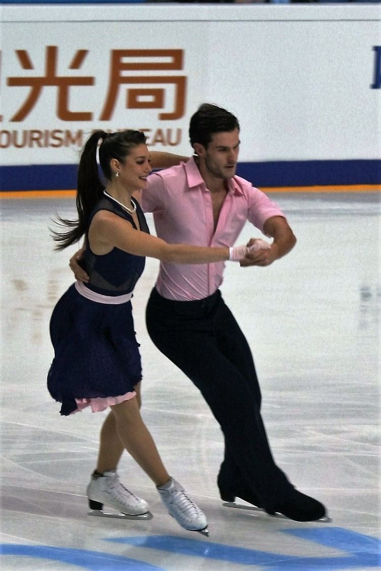 Laurence Fournier Beaudry and Nicolaj Sorensen performing a dance in the ice rink during the 2016 Rostelecom Cup in Russia. Laurence wearing a black and pink dress and white figure skate while Nicolaj wearing a pink shirt, black pants, and black figure skate