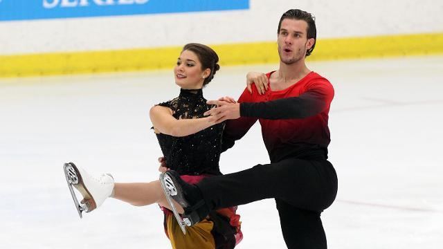 Laurence Fournier Beaudry together with her male partner Nikolaj Sørensen captured while performing a short dance during the official training at Trophee Bompard ISU Grand Prix at Patinoire Meriadeck Arena in France. Laurence, smiling with an all tied up hair, light makeup, and silver stud earrings, holding Nikolaj’s left hand and her left hand on Nikolaj's shoulder while raising her left leg 90 degrees in between, with her figure skates shoes and she's wearing a black lace neck sleeveless dress with a shade of red below her dress. Nikolaj, blow mouth look with a beard, holding Laurence’s right hand and his right hand on Laurence’s waist while raising his left leg 90 degrees in between with his figure skates shoes and he's wearing a shade of red long sleeve shirt and black pants.