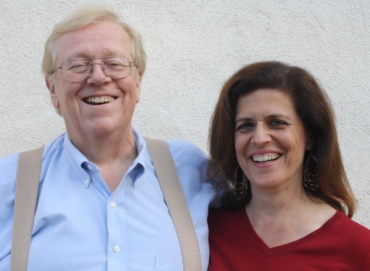 Leda Cosmides smiling and wearing a red blouse while John Tooby wearing blue long sleeves and eyeglasses
