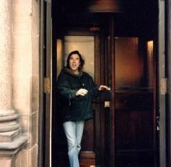 Lee Harris (drummer) wearing a black jacket and denim pants while standing in front of a door