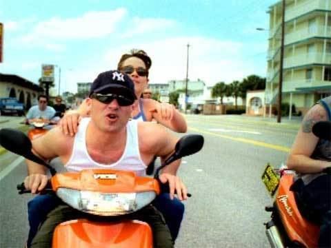 Marc Costanzo wearing a cap, sunglasses, a white sleeveless shirt, and driving an orange motorcycle with Sharon Costanzo riding at the back.