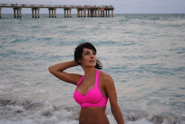 Lena Miro holding her hair and looking at something above while at the beach and wearing a pink swimsuit