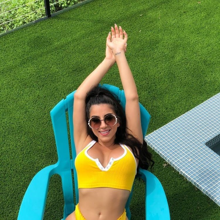 Lena The Plug smiling while sitting on a blue chair doing hands up, with long wavy hair, wearing sunglasses, and a yellow two-piece swimsuit.