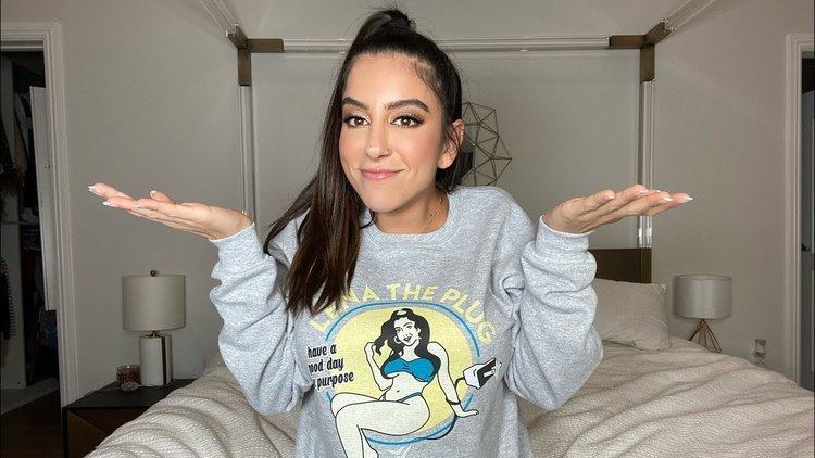 Lena The Plug smiling, doing an I don't know expression gesture while sitting on the bed, with long hair, and wearing a gray sweatshirt.