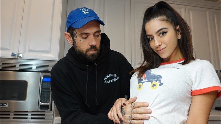 Lena The Plug smiling while Adam Grandmaison touching her tummy. Lena wearing a white shirt while Adam wearing a blue cap and black jacket.