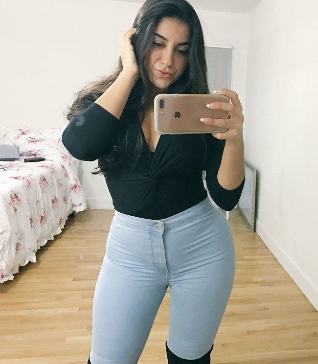 Lena The Plug doing pouty lips in front of a mirror while holding a phone, with long wavy hair, wearing a black long sleeves top, light blue pants, and black boots.