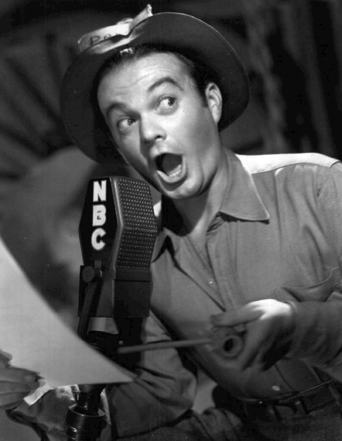 Leo Gorcey with a surprised face while holding a paper and a microphone, wearing a black hat, gray long sleeves, a belt, and gray pants.
