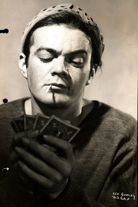 Leo Gorcey with a suspicious face while looking at the cards and smoking a cigarette, wearing a gray hat, and a black long sleeve shirt.