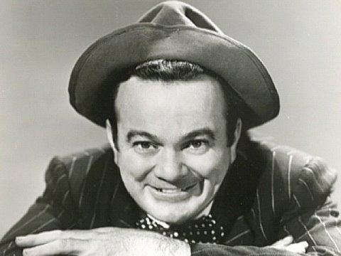 Leo Gorcey is smiling, wearing a black hat, a black stripe coat over white long sleeves, and a polka dots bow tie.