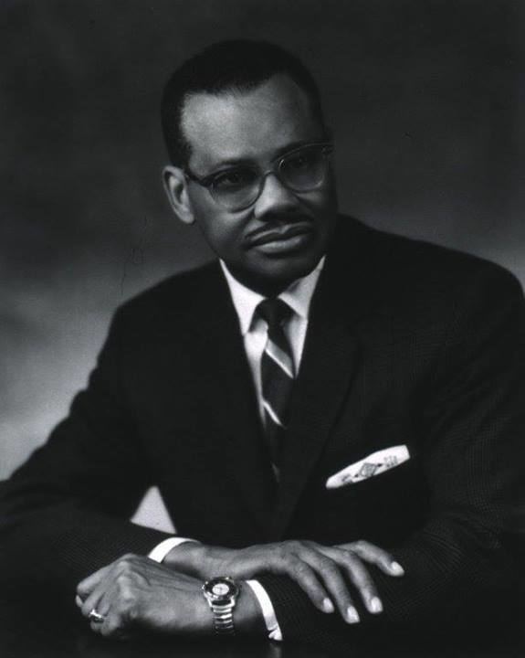 Leonidas Berry looking serious with a mustache with his right hand holding his left arm resting on the table, wearing a watch, a finger ring, eyeglasses, with a printed handkerchief in its pocket, and a white shirt with a necktie under a black coat.